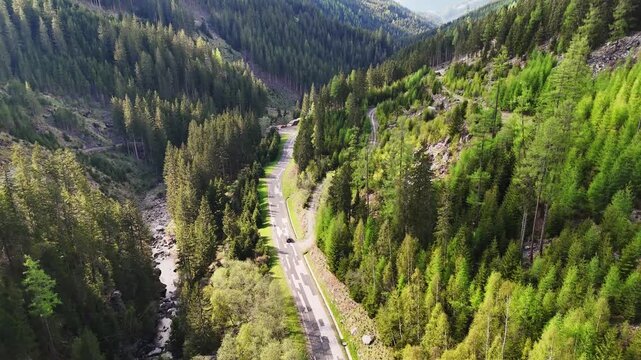 Aerial view of a winding road through a dense coniferous forest valley with a mountain stream and steep slopes in Murau, Styria, Austria.