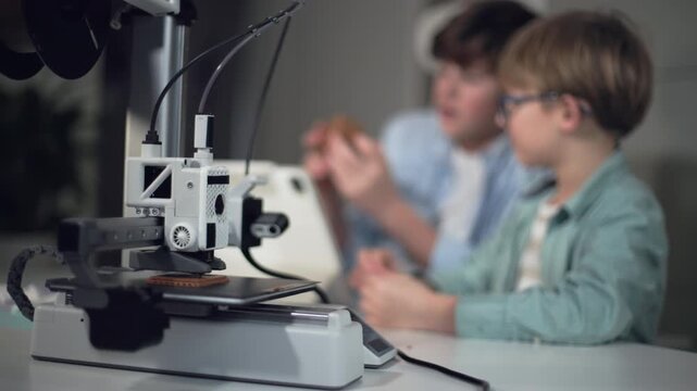 The school boys at a table with a working 3D printer, observing and operating it