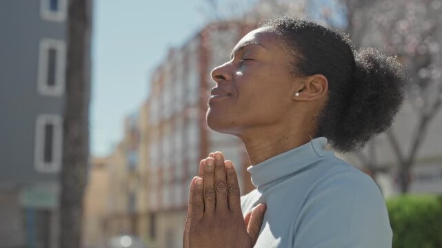 Woman praying with hands pressed together and closed eyes, looking upward on a sunny urban street beside apartment buildings and trees; serenity.