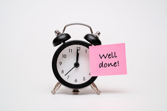 Black alarm clock with a pink sticky note text "WELL DONE!". Concept of productivity, timely success, morning achievement, and positive reinforcement