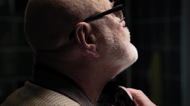 A silver-haired man shaves with a trimmer in front of a mirror in the bathroom