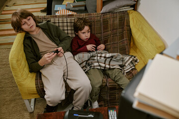 Obraz premium High angle view of Caucasian kids playing video game on couch, sharing screen time at home and showing low income family lifestyle. Useful for parenting, poverty, childhood content
