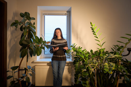 Woman in patterned sweater reading a book by the window surrounded by houseplants