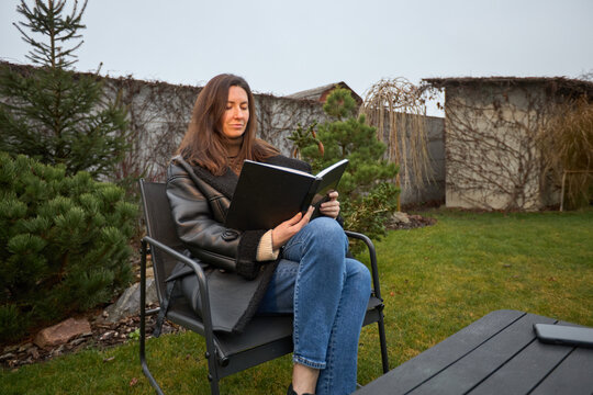 Woman in warm coat reading a book outdoors in a garden setting