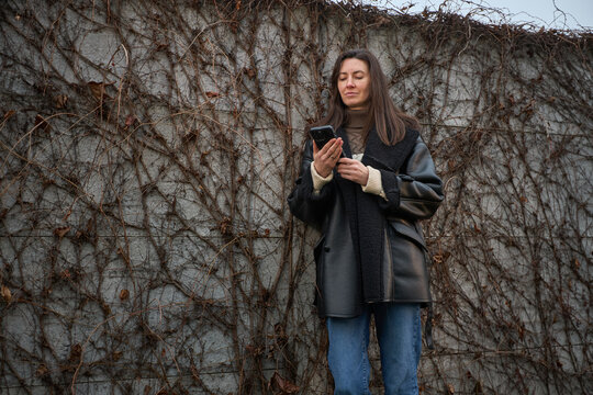 Woman in stylish winter coat using smartphone against a textured wall covered in dry vines