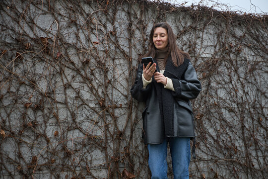 Woman in warm winter coat using smartphone against a textured wall covered in dry vines