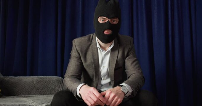 Scammer in balaclava demands money while sitting on couch in dark room. Man pinches fingers forward pointing ahead to pressure victim during fraud call moment