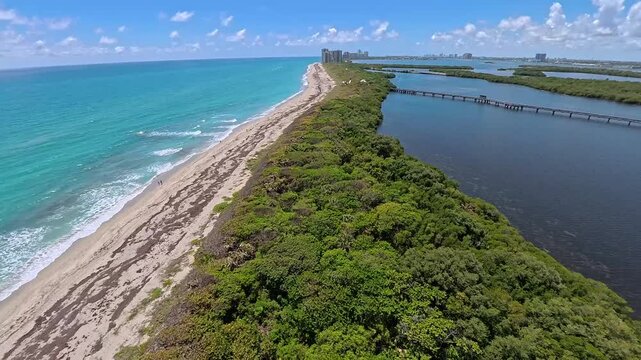 Aerial view of McArthur Beach with its sandy coastline, turquoise ocean waves, lush mangroves, and a long wooden bridge in North Palm Beach, Florida, United States.