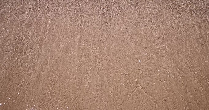 Slow motion footage of gentle sea water washing onto a sandy beach with soft foam and textured wet sand creating a peaceful coastal background scene with natural seaside detail in a relaxing setting