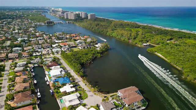 Aerial view of luxury waterfront homes along winding canals with boats, lush greenery, and high-rise buildings near the ocean coast in Boca Raton, Florida, United States.