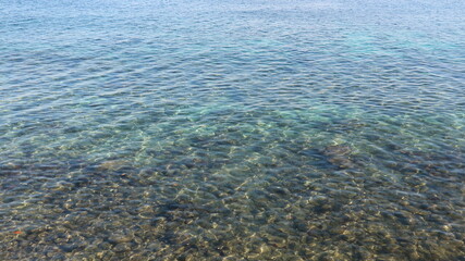 Obraz premium Clear water gently ripples over rocks below. Shallow sea reveals a bed of smooth stones. Light dances across the water's surface. Ideal for tranquil nature or travel visuals. No people, no animals
