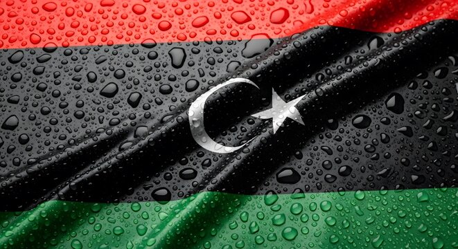 The national flag of Libya, featuring red, black, and green stripes with a white crescent and star, is covered in numerous glistening water droplets.