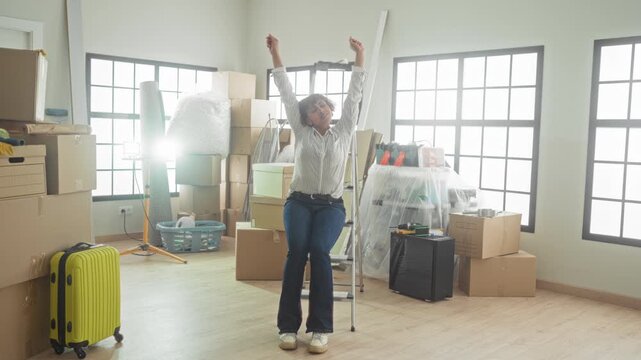 Woman raising arms over packed boxes and yellow suitcase in building during move; relief beginning renewal.