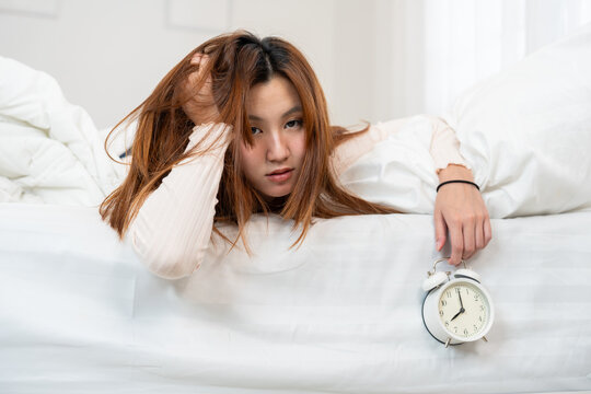 Tired young Asian woman messy hair lying on bed holding alarm clock at 8 AM, feeling exhausted and suffering from insomnia or sleep deprivation in white bedroom. Morning laziness concept.