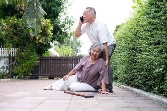 Senior Asian woman fallen on ground with knee injury while husband uses smartphone to call emergency services. Ideal for healthcare, elderly safety, and medical insurance marketing themes