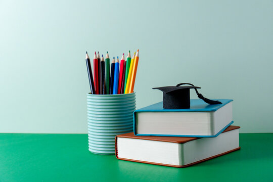 A miniature graduation cap on a stack of books next to a container of colored pencils. Graduation, academic success, higher education, school readiness, and educational milestones concept.