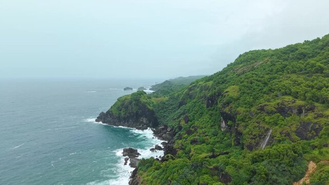 Aerial view of Bukit Pengilon featuring lush green cliffs and a small waterfall cascading towards the sea in Kecamatan Tepus, Daerah Istimewa Yogya, Indonesia.
