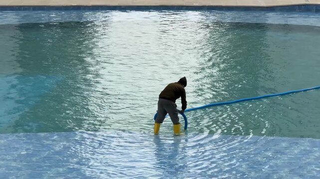 Person cleaning outdoor swimming pool with long blue hose, wearing yellow boots on a clear day