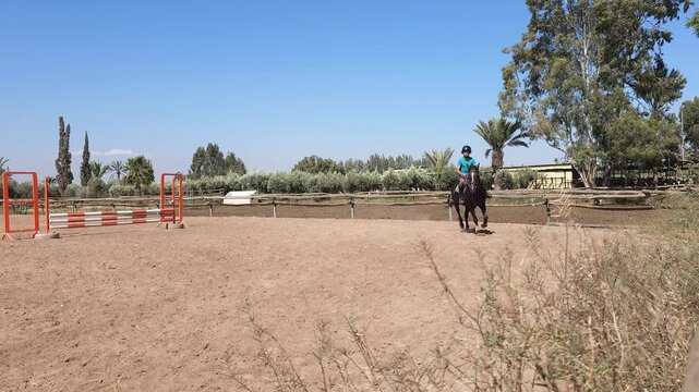 Young rider practicing horseback riding on a chestnut horse in an outdoor riding arena surrounded by greenery. Child equestrian wearing a safety helmet during riding lesson at equestrian club on a sun
