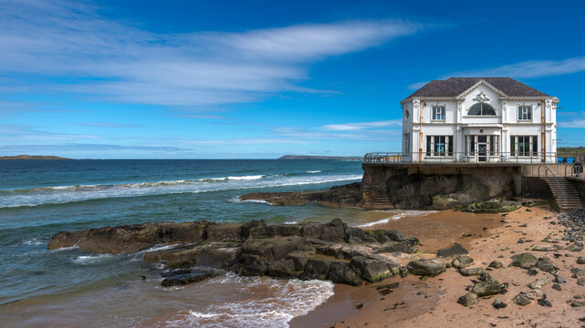 Historic white building on the rocky coast of Portrush, Northern Ireland. Atlantic Ocean waves hitting the beach under a blue summer sky. Iconic coastal architecture and travel destination.