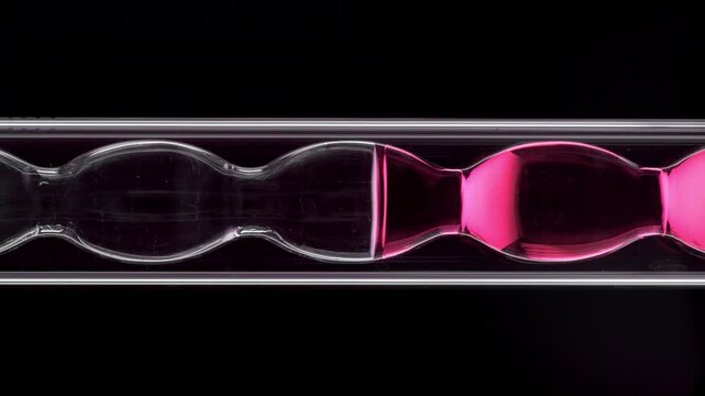 Bold vertical close-up of air bubbles floating through transparent rose liquid as it fills and drains from verically installed bulb laboratory condenser on solid black surface