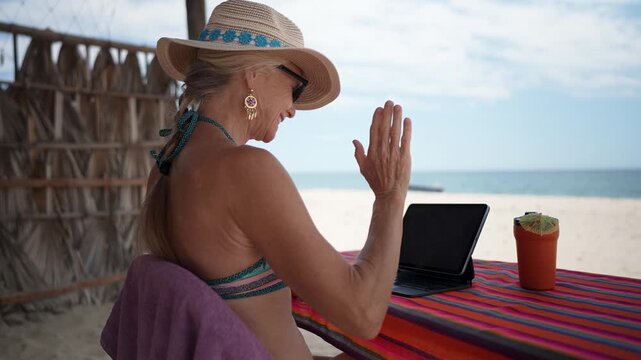 Mature woman has video call on her computer while sitting at a beachside table during her vacation, expressing happy emotions under the sun.