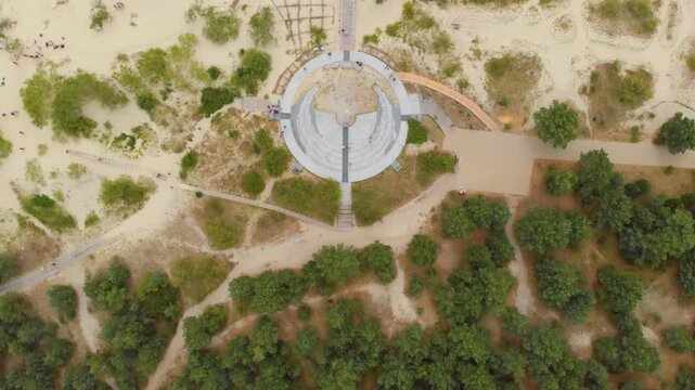 Drone footage capturing a top down view of the modern Nida Sun Clock monument, located on a sandy hill amidst green forests on the Curonian Spit, Lithuania.