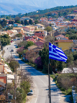 Aerial view of Greek flag waving proudly over the picturesque coastal town of Acharavi in Corfu Greece with traditional houses road and distant mountain landscape under blue sky.