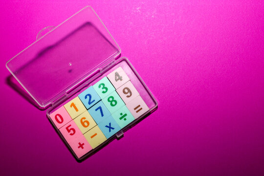 A children's math set with colorful numbers and signs is neatly arranged inside an open transparent plastic box. The learning kit is located on a bright purple-pink background with free space for text