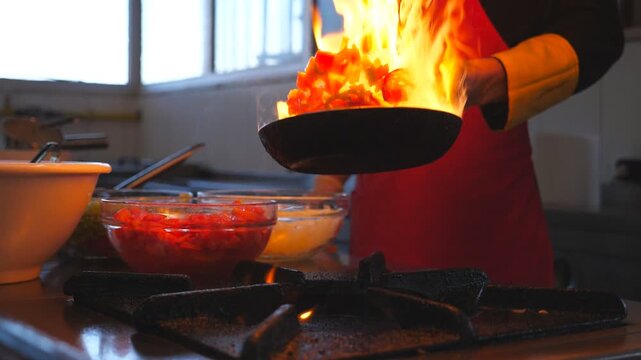 Unrecognizable chef cooking tasty flambe style dish on pan for visitors of restaurant. Professional cook tossing and frying vegetables in flaming skillet on stove. Side view Slow motion Close up