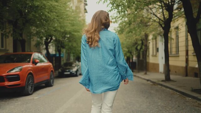 A playful young woman in yellow sunglasses turns back to smile at the camera while walking down a cozy city street. Urban lifestyle, casual clothing, cheerful mood, female portrait, summer travel.