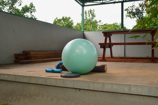 An angled perspective from the front-right captures a teal stability ball, a kinetic twister tool, and an expander trimmer band on a pavilion floor amidst a green orchard.