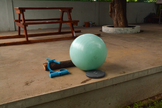 A high-angle view looking down from the front-left onto a teal workout ball, a waist twister, and a body trimmer on a building floor in a park rest station.