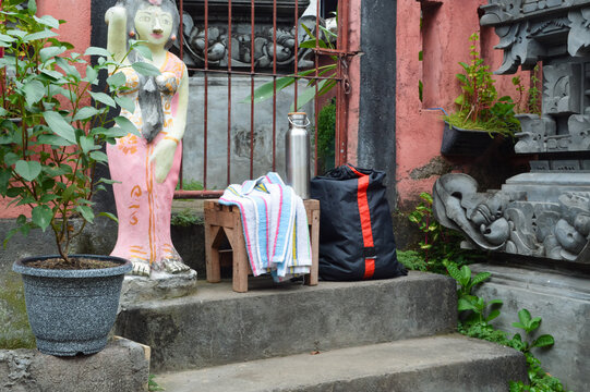 Looking from a front-left angle, a black athletic backpack, a stainless steel drink bottle, and a white towel rest on stone steps near a traditional Balinese shrine structure.