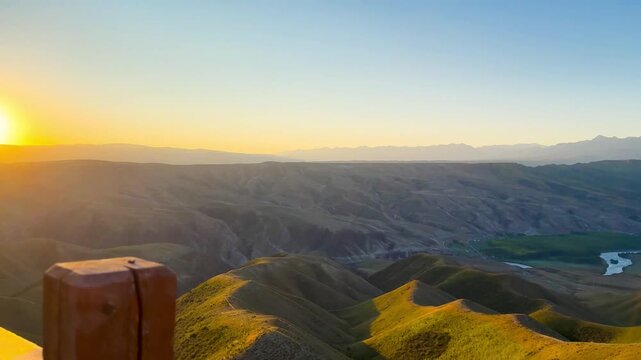 A panning video of mountains with beautifully overlapping layers of green and golden yellow at dawn in Xinjiang, China. The sun is rising with lens flares and warm tones.