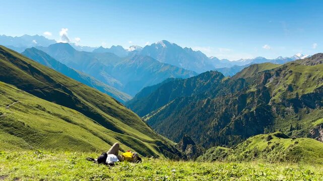 A person lies down on the green grass on the Tian Shan Mountains, China. It has views of mountain peaks and pine forests as a backdrop.
