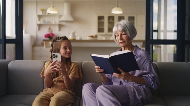 Senior woman reading a book while granddaughter plays on phone, generation gap