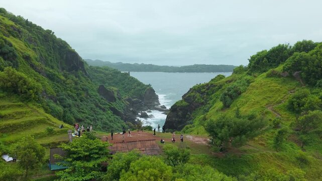 Aerial view of Bukit Pengilon with people standing on lush green hills overlooking the rocky coastline and Indian Ocean in Kecamatan Tepus, Daerah Istimewa Yogya, Indonesia.