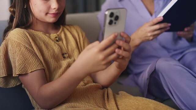 Young girl scrolling on smartphone while her grandma reads a book nearby
