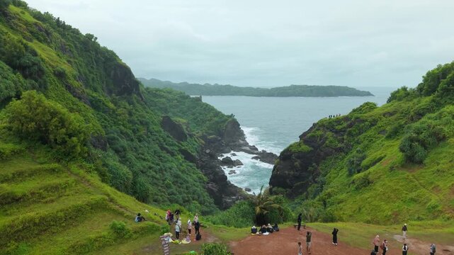 Aerial view of Bukit Pengilon featuring lush green cliffs, crashing waves on the rocky shore, and people on the edge in Kecamatan Tepus, Daerah Istimewa Yogya, Indonesia.