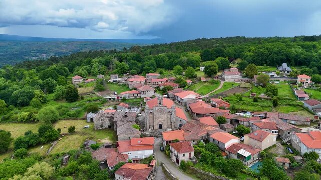 Aerial view of Iglesia de Santa Marina de Augas Santas surrounded by red-tiled roofs and lush green forests under a cloudy sky in Augas Santas, Ourense, Spain.