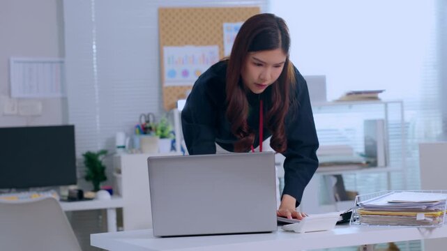 Young Asian businesswoman working at office desk with laptop computer reviewing document folder then showing serious focused expression discovering unexpected corporate news preparing presentation