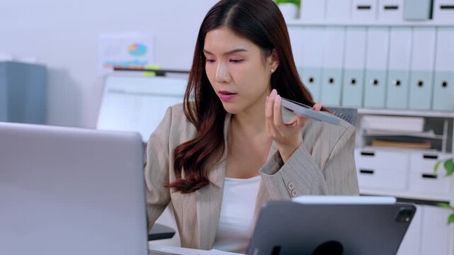 Frustrated Asian business woman holding smartphone speaking loudly complaining about bad news on laptop computer screen looking angry stressed at modern corporate office desk with tablet folders