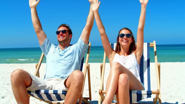 Smiling caucasian man and woman in beachwear raise arms joyfully from chairs on sand beach, turquoise ocean. Aspirational travel commercial aesthetic