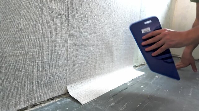 Person smoothing textured wallpaper along interior wall baseboard