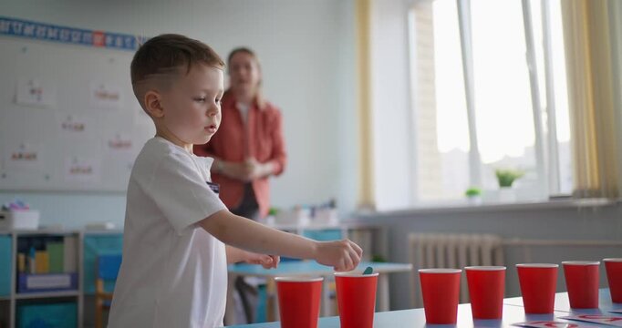 Young boy practicing a classroom cup sorting activity with red cups while a teacher observes in a bright early learning room