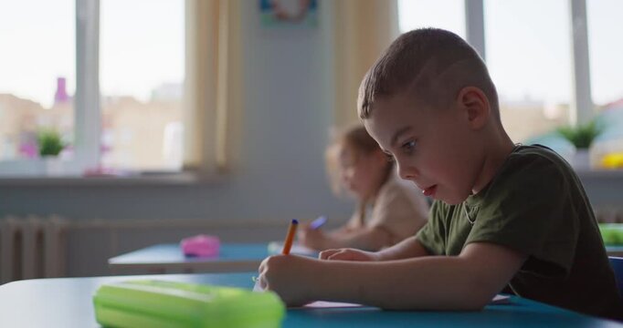 Focused young boy writing with a pencil at a classroom desk during a quiet lesson, with soft window light and another child studying nearby