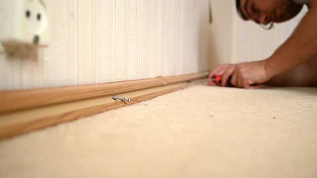 Man removing wooden baseboard trim along interior wall on carpet
