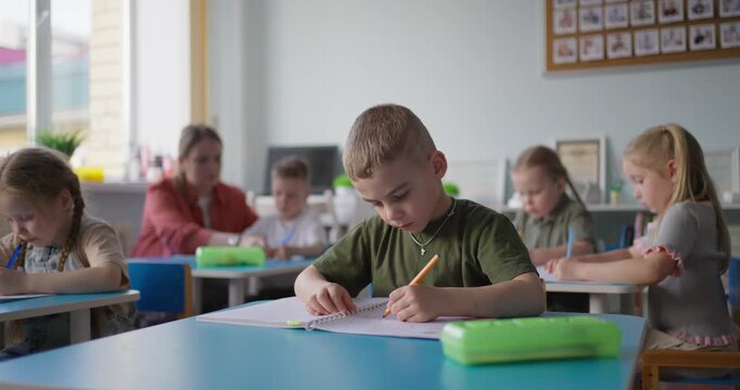 Young children concentrate on writing with pencils at blue desks in a bright classroom while a teacher supervises a quiet learning activity