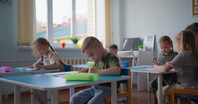 Young students concentrate on writing at individual desks in a bright classroom while a teacher guides a quiet elementary lesson
